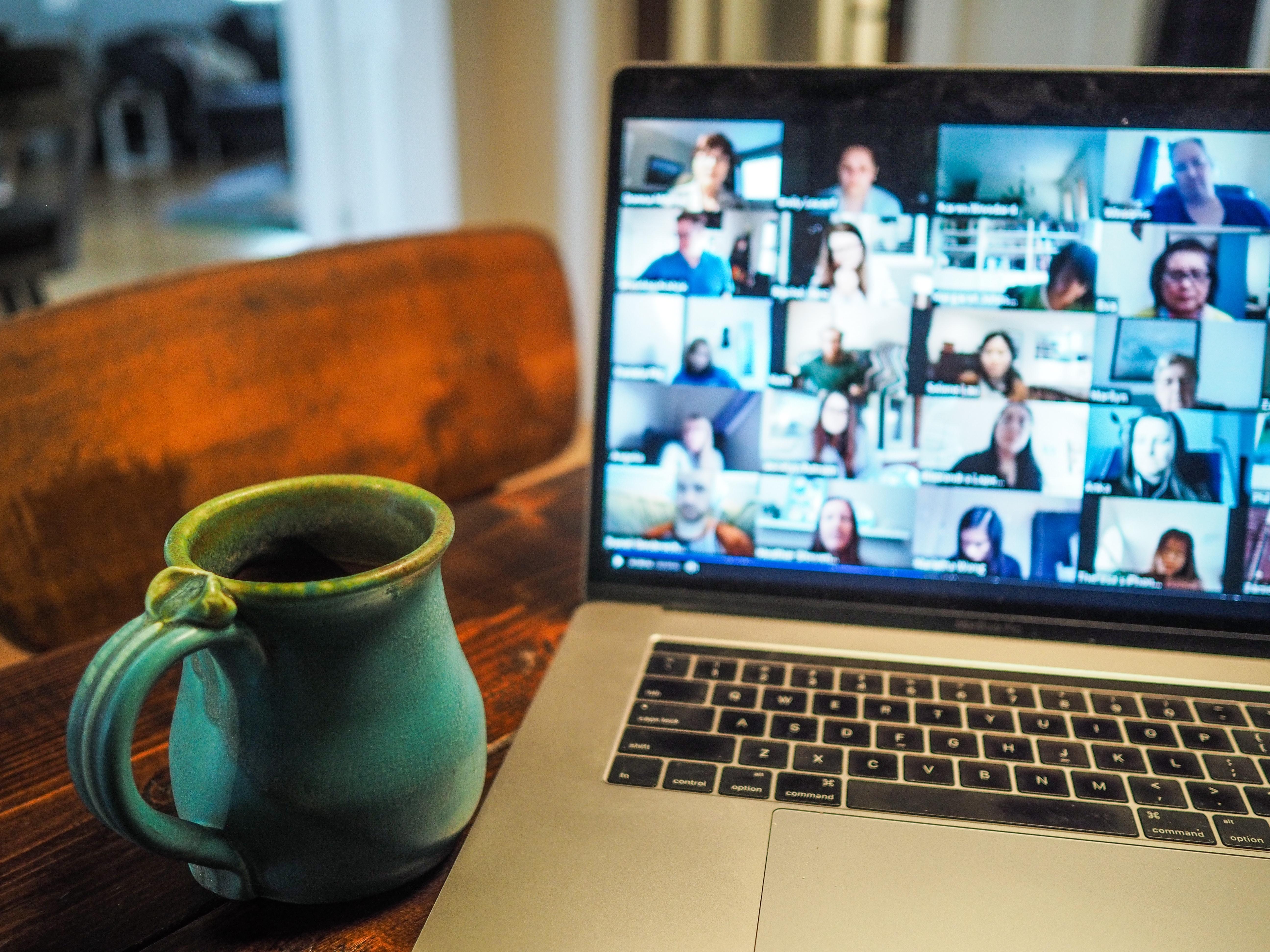 Photo of a laptop showing a video call in progress, there are lots of different people on the screen. There is a green mug in the left hand corner. 